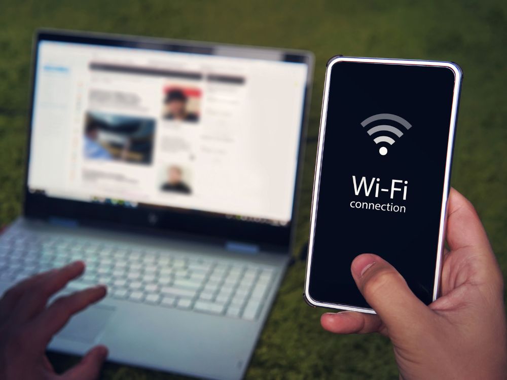 Pessoa segurando celular com tela de conexão Wi-Fi ao lado de notebook. Cena ilustrando uso de dispositivos conectados, ideal para o tema Descubra o que causa interferência no Wi-Fi.