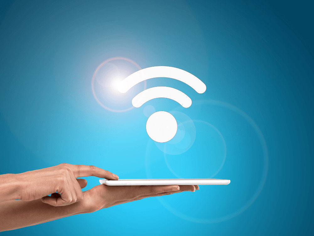 Mão segurando um tablet abaixo do símbolo de Wi-Fi, representando acesso à internet por conexão sem cabos.