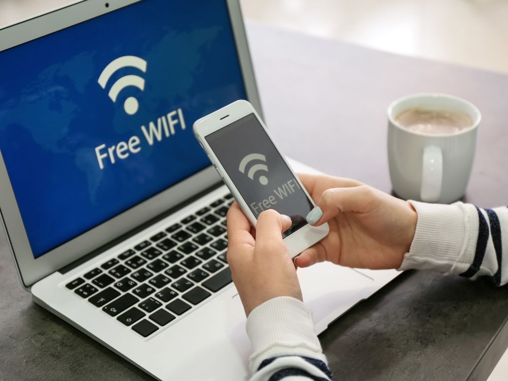 Usuário utilizando smartphone com mensagem de Wi-Fi gratuito em frente a notebook. Imagem que representa ambientes compartilhados e ajuda a contextualizar Descubra o que causa interferência no Wi-Fi.